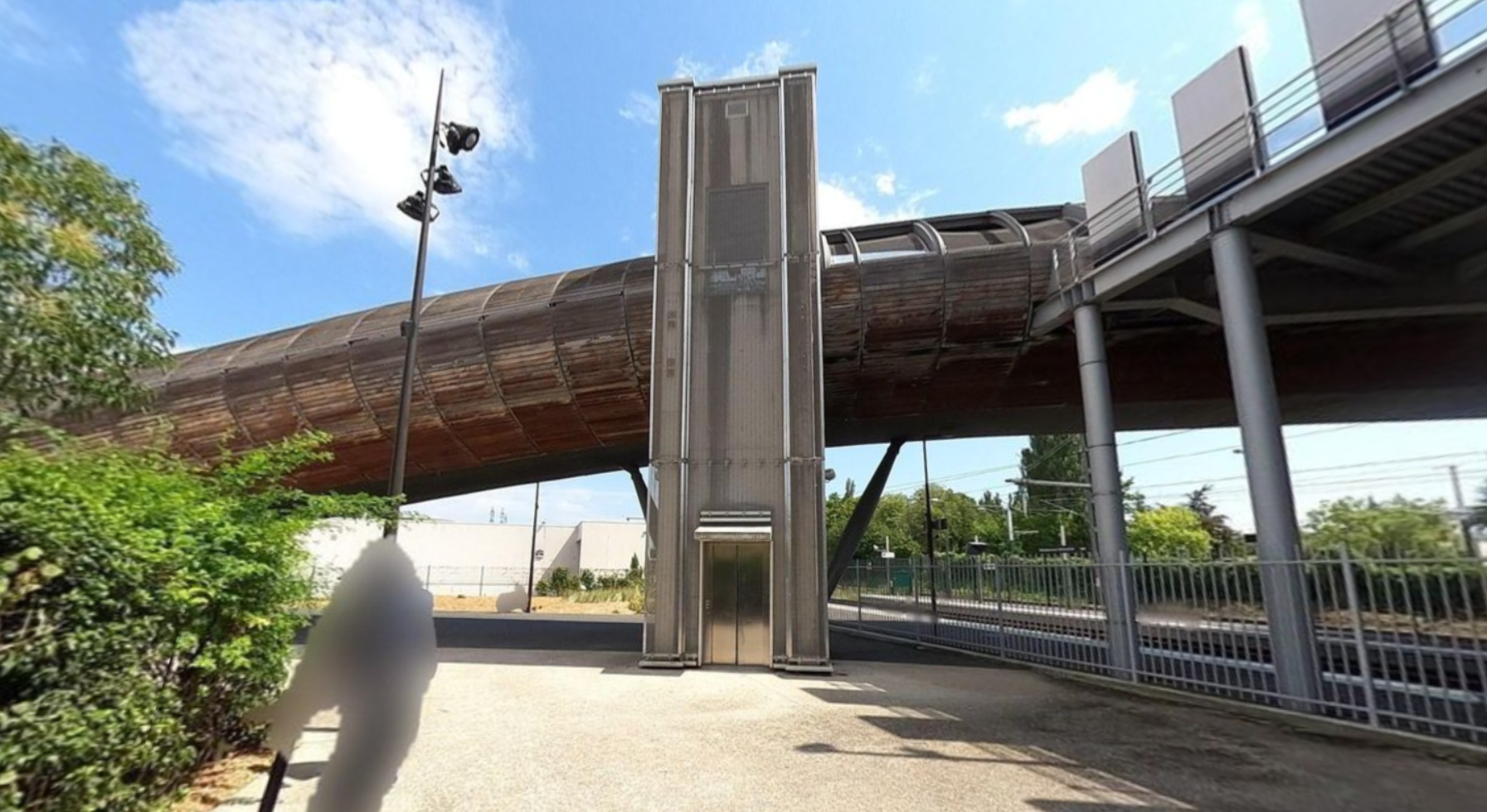 photo : on voit un ascenseur qui permet d'accéder à une passerelle qui passe par dessus des voies ferrées